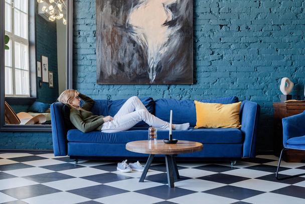Young caucasian woman in green sweater and white jeans lying on sofa in stylish blue living room, using mobile - Foto, afbeelding