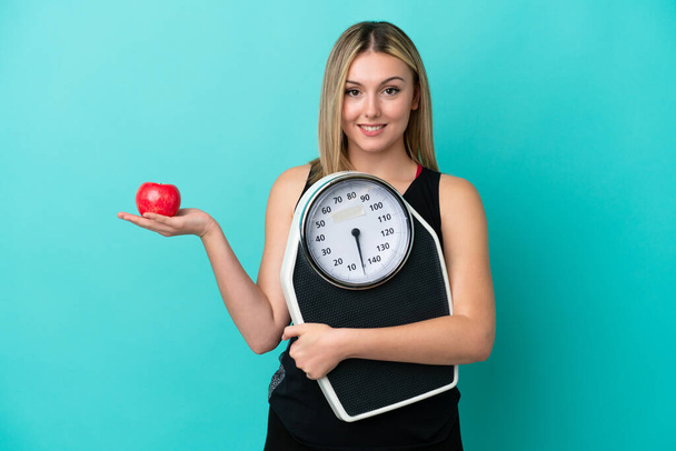 Young caucasian woman isolated on blue background with weighing machine and with an apple - Photo, Image
