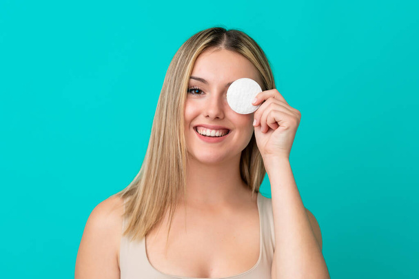 Young caucasian woman isolated on blue background with cotton pad for removing makeup from her face and smiling - Foto, immagini