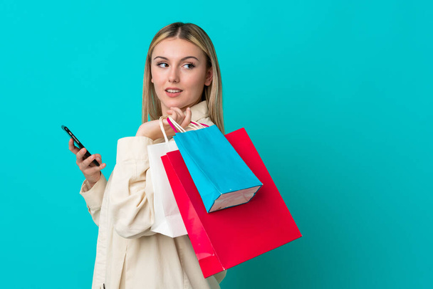Mulher caucasiana jovem isolado no fundo azul segurando sacos de compras e um telefone celular - Foto, Imagem