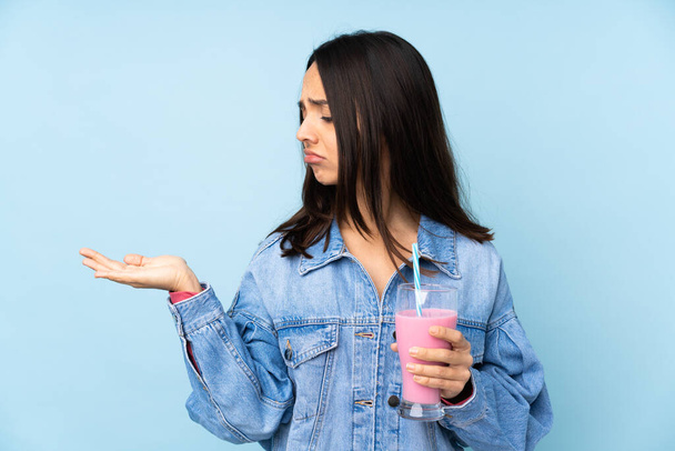 Jeune femme avec milkshake fraise sur fond bleu isolé tenant copyspace avec des doutes - Photo, image
