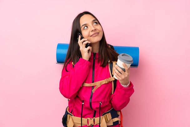Młoda dziewczyna alpinistka z dużym plecakiem na odizolowanym różowym tle trzymając kawę do zabrania i mobilny - Zdjęcie, obraz