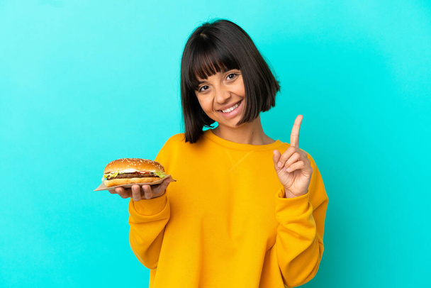 Młoda brunetka trzyma hamburgera na odosobnionym tle pokazując i podnosząc palec - Zdjęcie, obraz