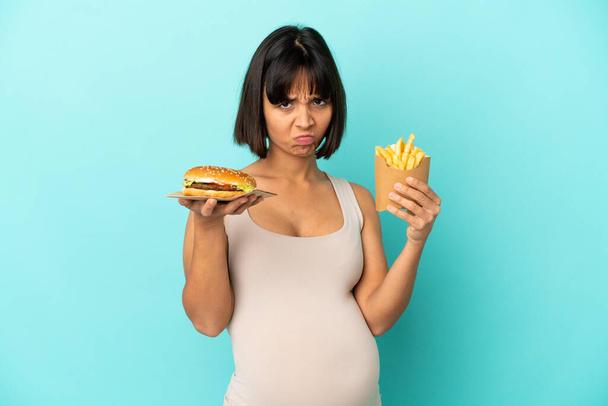 Молода вагітна жінка тримає бургер і смажені чіпси на ізольованому синьому фоні
 - Фото, зображення