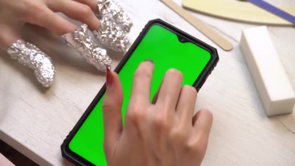 Een meisje gebruikt tijdens een manicure een smartphone met een groen scherm. Zelfgemaakte manicure service. Manicure verft nagels met roze gellak. Gemanicuurde rode nagels. Nagellak aanbrengen. - Video