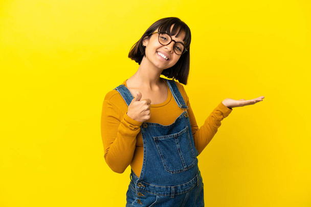 Jonge zwangere vrouw over geïsoleerde gele achtergrond houden copyspace imaginaire op de palm om een advertentie in te voegen en met duimen omhoog - Foto, afbeelding