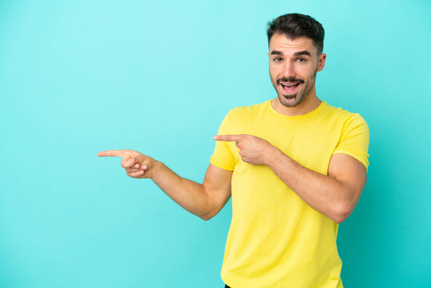 Young caucasian man isolated on blue background surprised and pointing side - Foto, imagen