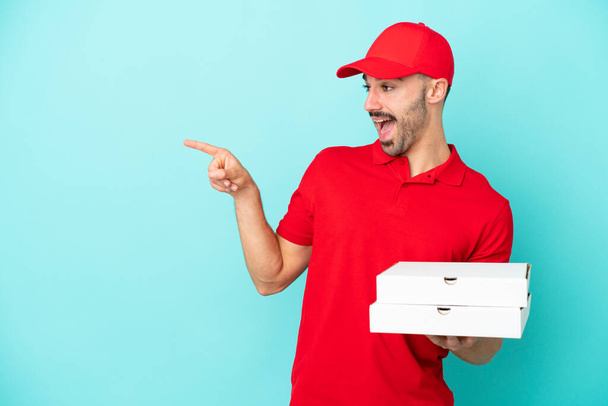 Lieferkaukasier nimmt Pizzakartons isoliert auf blauem Hintergrund entgegen und zeigt mit dem Finger zur Seite und präsentiert ein Produkt - Foto, Bild