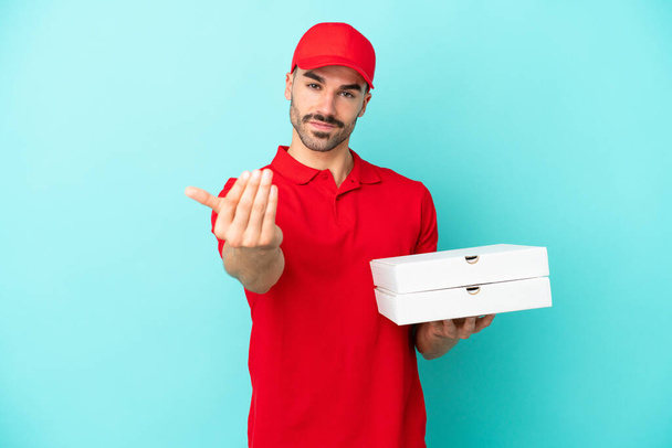 Lieferkaukasier, der auf blauem Hintergrund isoliert Pizzakartons aufhebt, die einladen, mit der Hand zu kommen. Glücklich, dass du gekommen bist - Foto, Bild