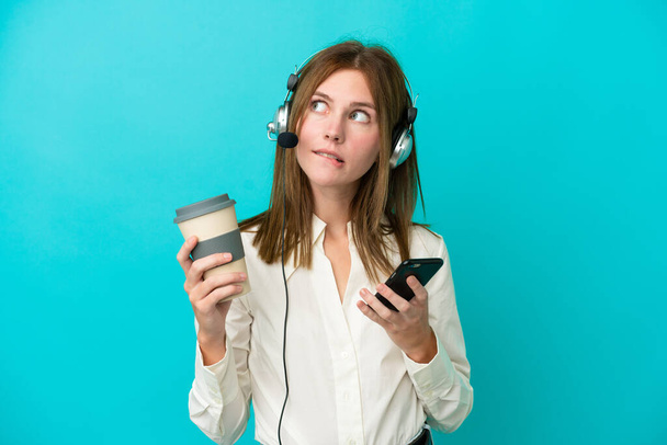 Telemarketer Russian woman working with a headset isolated on blue background holding coffee to take away and a mobile while thinking something - Фото, изображение