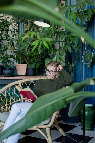 Tiempo libre. Joven mujer rubia sentada en silla cómoda leyendo un libro rodeado de plantas - Foto, imagen