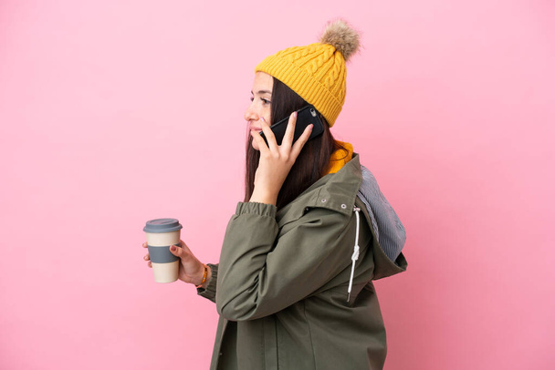 Young Ukrainian woman wearing winter jacket isolated on pink background holding coffee to take away and a mobile - Фото, зображення