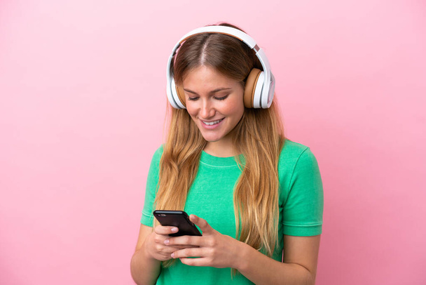 Genç sarışın kadın pembe arka planda izole edilmiş müzik dinliyor ve cep telefonu arıyor. - Fotoğraf, Görsel