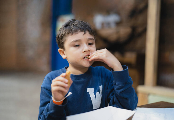 Голодний хлопчик їсть палицю Churros, Шкільний хлопчик перекушує після обіду в шкільному кафе, Здорова дитина сидить у кав'ярні їсть їжу
. - Фото, зображення