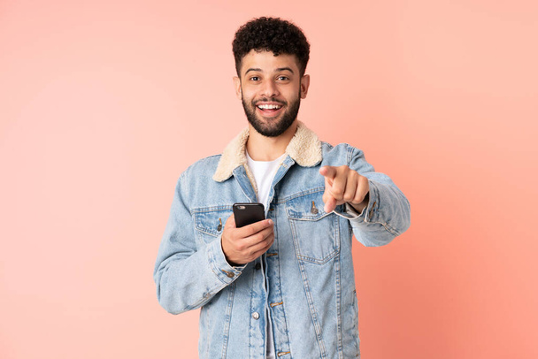 Jeune homme marocain utilisant un téléphone portable isolé sur fond rose surpris et pointant devant - Photo, image