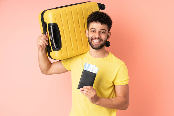 Faslı genç bir adam pembe arka planda izole edilmiş, tatilde bavul ve pasaportla. - Fotoğraf, Görsel