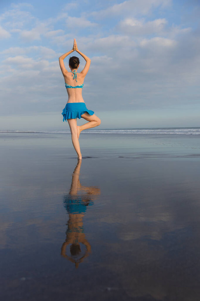 Yoga pose. Vrikshasana asana. Young woman practicing tree pose at the beach near the ocean during sunset. Arms raised in namaste mudra. Outdoot yoga. Water reflection. View from back. Copy space. Bali - Photo, Image