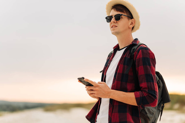 Happy young man in sunglasses and hat uses his cell phone enjoying a weekend in the mountains. Traveler standing on top of a mountain at sunset and using a mobile phone - Fotografie, Obrázek