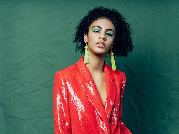 красивая женщина в красной куртке ювелирные изделия яркий макияж зеленый фон - Фото, изображение