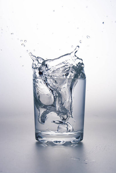 Wasserglas mit Spritzer auf hellgrauer Oberfläche und weißem Hintergrund. - Foto, Bild