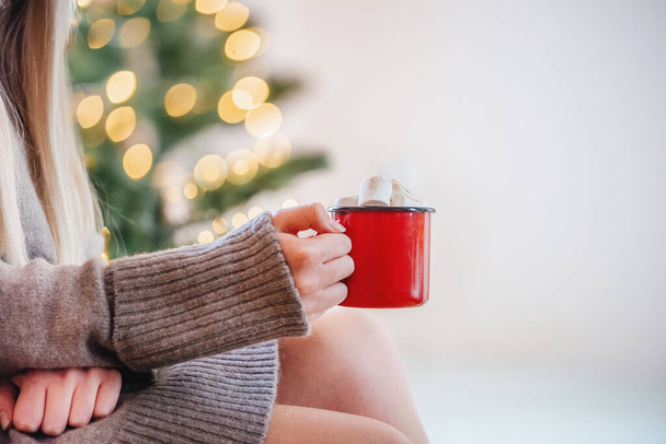 Hände, die eine rote Tasse heiße Schokolade mit Marshmallows auf Weihnachtsbaumbeleuchtung Bokeh Hintergrund - Foto, Bild