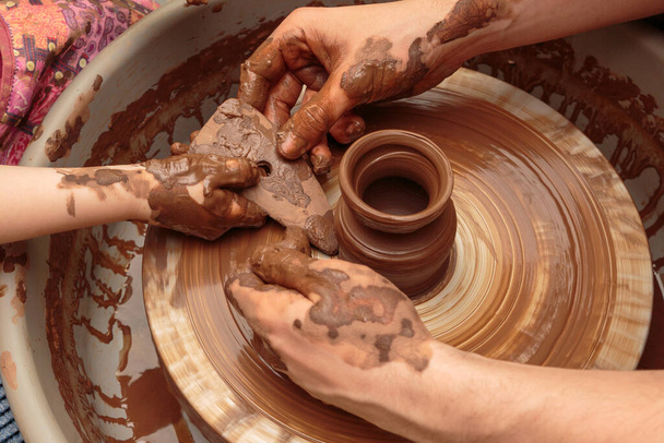 子供は陶芸家の車輪の上に鍋を粘土から成形します。大人は子供が粘土で作業するのを助ける. - 写真・画像