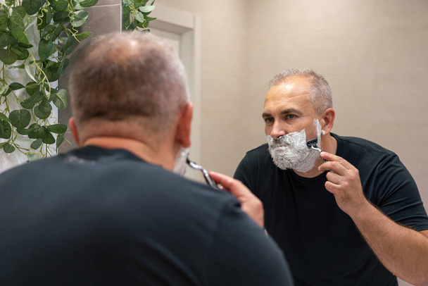 Hombre de mediana edad cortando su barba y bigote de pie en el baño y mirando en un espejo - Foto, imagen