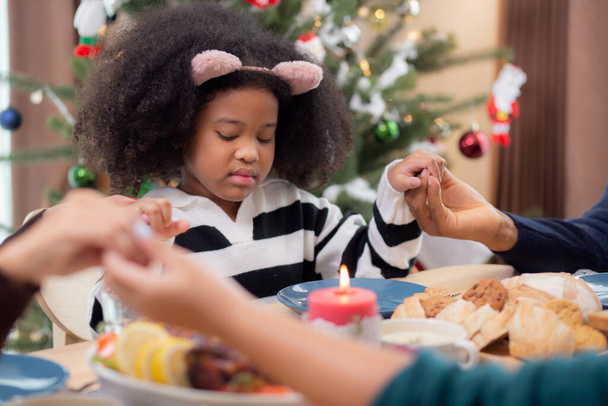 幸せなアフリカの家族が手を取り合って感謝祭の日に夕食を祈る一緒に、陽気なクリスマス、フードテーブルでのお祝いとお祝い、家族の関係、屋内、新年とクリスマス. - 写真・画像