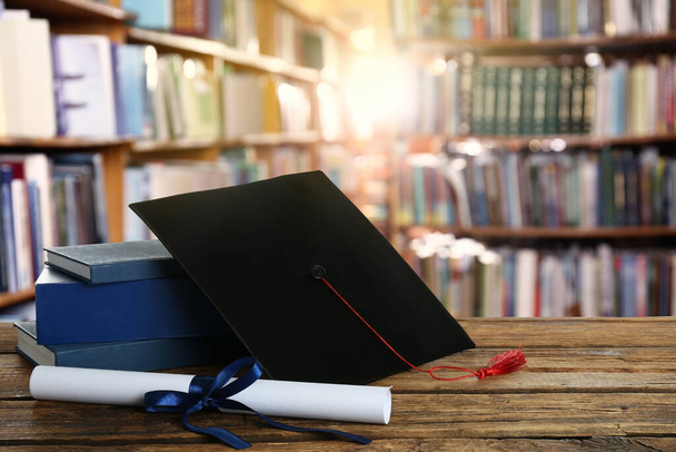 Graduation hat, books and diploma on wooden table in library - Foto, Bild