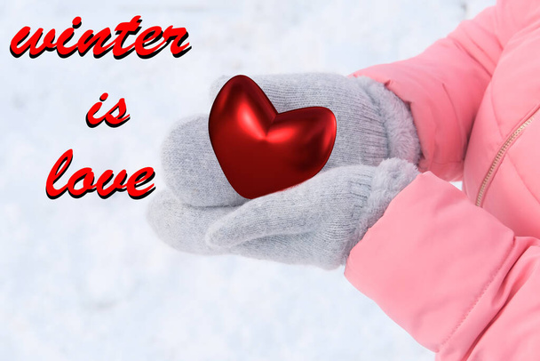 girl in a pink jacket holding a red heart model, a symbol of love, gray woolen mittens in her hands, winter background and white snow, concept of confession, Valentine's Day - Photo, image