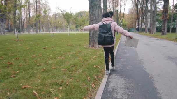 Zpět pohled školačka chodit na obrubníku holčička s černým batohem udržet rovnováhu dítě jít do školy držení knihy v ruce neznámé dítě chůze v městském parku po studiu africký americký žák hrát sám - Záběry, video