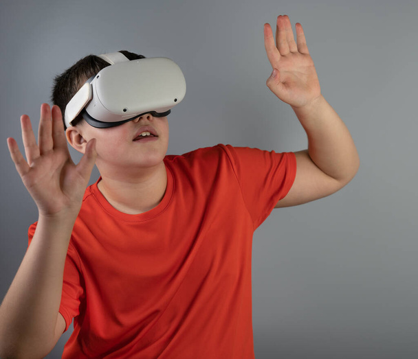 Ένας έφηβος με γυαλιά-viar κοιτάζει ψηλά και σήκωσε τα χέρια του, ενώ παίζει ένα παιχνίδι. Ένα αγόρι με κόκκινο μπλουζάκι σε γκρι φόντο. Εικονική πραγματικότητα - Φωτογραφία, εικόνα