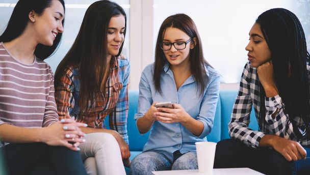 Group of glad girlfriends in casual clothes sharing smartphone while watching video in social network spending free time together in light room - Photo, image