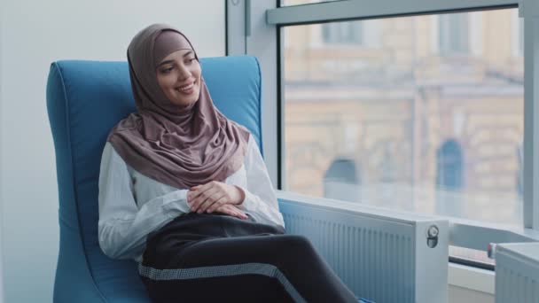 Lachend dromerig Arabisch meisje in hijab kijkend uit raam zittend in fauteuil wachtend op examen of interview resultaten. Kandidaat bij recruit agent kantoor verwacht een ontmoeting met de werkgever voor het inhuren - Video
