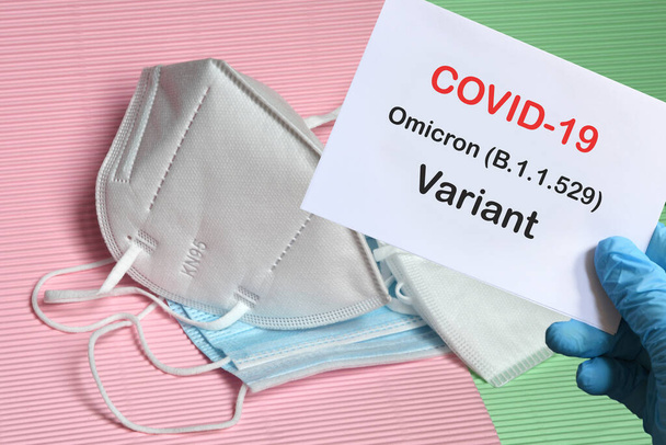 Doktorun elinde beyaz kağıtlı mavi eldiven ve arka planda çeşitli koruma maskeleri olan Covid-19 Omicron Variant mesajı var. COVID-19 omikron varyant türü koruma kavramı. - Fotoğraf, Görsel