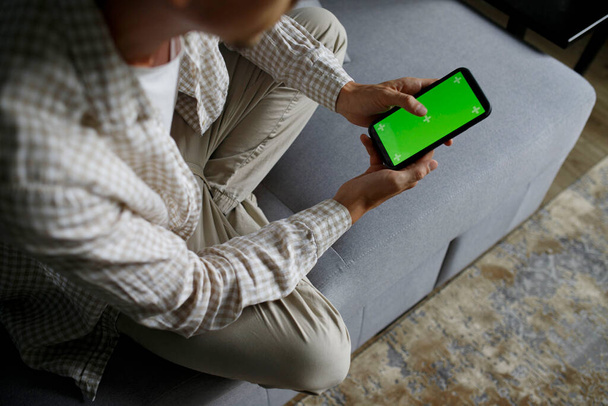 Close up of a man using mobile smart phone with green screen sitting in living room on sofa - Valokuva, kuva