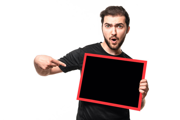 Парень с открытым ртом указывает пальцем пространство для текста или объявления на плакате. Удивленный мужчина в черных футболках показывает рекламу в студии - Фото, изображение