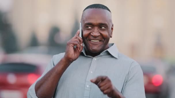 Hombre de negocios afroamericano parado en la calle en el centro de la ciudad sosteniendo un teléfono inteligente, hombre negro profesional divirtiéndose, hablando emocionalmente por teléfono con un amigo o pareja, hombre anciano étnico al aire libre - Imágenes, Vídeo