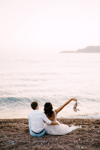 Жених обнимает невесту, сидя на песчаном пляже у моря. Вид сзади - Фото, изображение