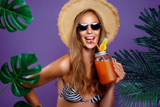 Summer fashion portrait of happy cheerful beautiful woman who drinking fruit cocktail, wearing trendy sunglasses, straw hat while stands near tropical plants isolated on purple studio background  - Fotoğraf, Görsel