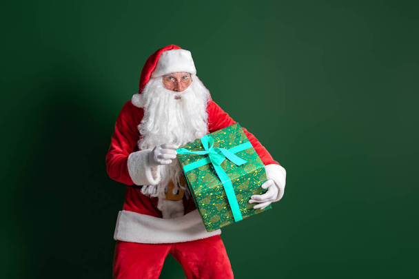 Santa Claus with long beard holds huge green gift box on green studio background while looking at camera - Fotoğraf, Görsel