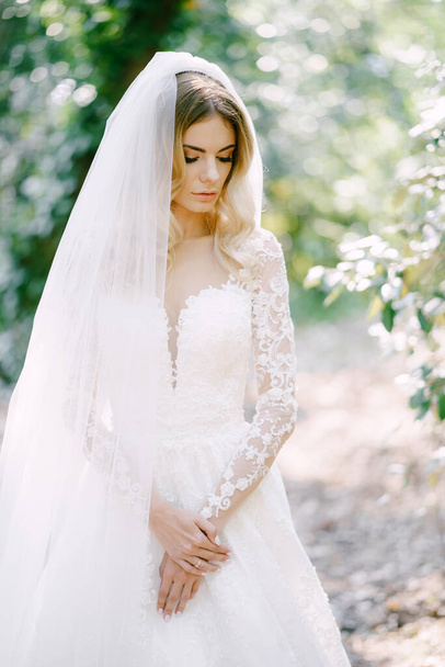 Mariée dans une robe en dentelle blanche se tient sur le fond des arbres avec ses yeux vers le bas. Photo de haute qualité - Photo, image