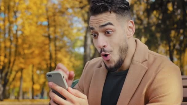 Nadšený hispánec čtení dobré zprávy na smartphone sedí na lavičce v podzimním parku mladý muž šťastně překvapen, že obdrží zprávu nadšený muž vítěz slaví vítězství držení telefonu venku - Záběry, video