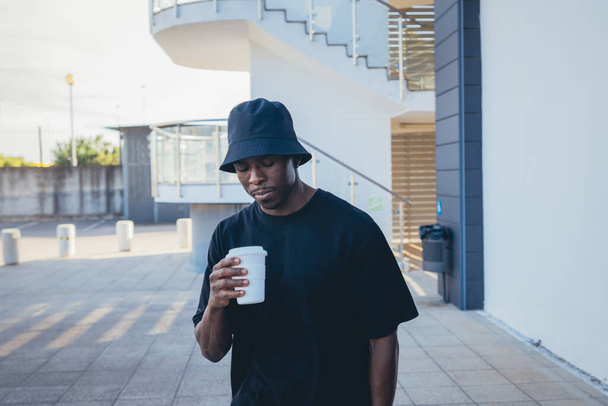 A young black man in street-style clothes holding his takeaway drink in front of a buildi - Photo, image