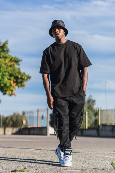 A vertical shot of a young black man in street-style clothes walking outdoors on a sunny day - Foto, immagini