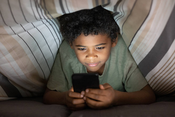 Niño negro lindo con Smartphone debajo del edredón - Foto, imagen