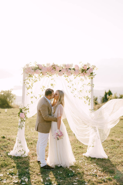 Gli sposi abbracciano e si baciano vicino all'arco nuziale, decorato con fiori delicati e tulle bianco  - Foto, immagini