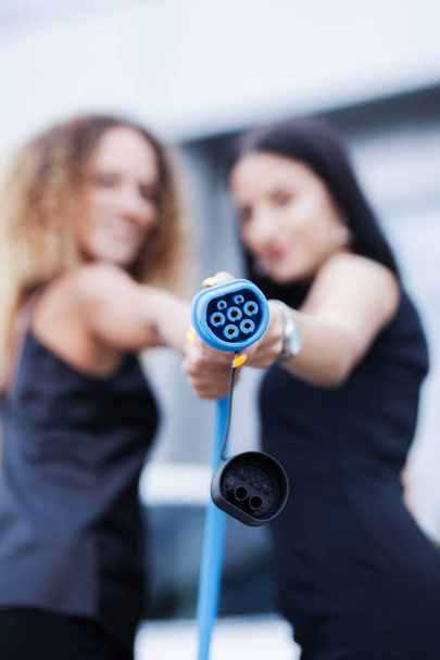 Two beautiful businesswomen in black outfut posing in front of an electric car. Girls holding blue charging cable. Shallow depth of field. Selective focus. - Foto, Imagen