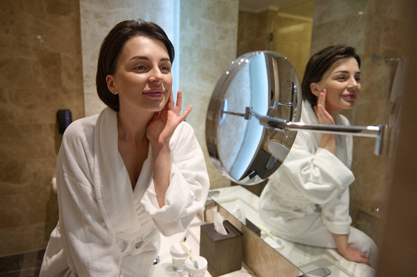 Привлекательная женщина в белом халате, улыбается, глядя на зеркальное отражение, трогает и делает антивозрастной сглаживающий массаж лица после нанесения увлажняющего крема на лицо. Концепция ухода за кожей - Фото, изображение