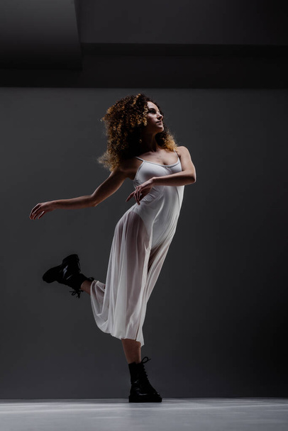 Девушка с вьющимися волосами, позирующая в балете. Балерина в белом платье и черных сапогах. - Фото, изображение